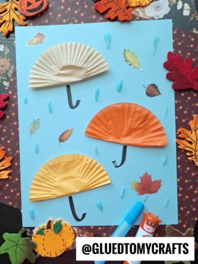 A blue paper displays three umbrella shapes made from folded cupcake liners with drawn handles. Blue raindrops, felt leaves, and craft supplies surround the artwork. The handle @GLUEDTOMYCRAFTS is at the bottom.
