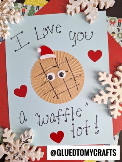 A handmade cupcake liner card craft featuring a paper waffle with googly eyes and a Santa hat, surrounded by hearts and snowflakes. The text reads “I love you a ‘waffle’ lot!” with “@GLUEDTOMYCRAFTS” at the bottom.