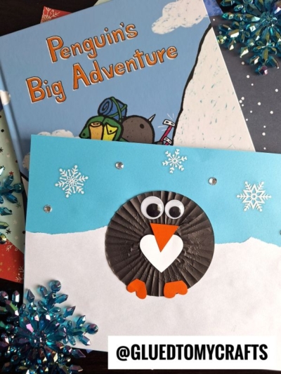 A handmade penguin card with a cupcake liner body, googly eyes, heart-shaped beak, and orange feet sits on a snowy background. Books and blue snowflake decorations are nearby. Text reads @GLUEDTOMYCRAFTS.
