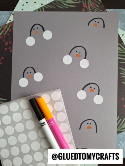 Gray paper with drawn penguin faces, each made from two white circular stickers for cheeks, like simple coding label crafts. Eyes and an orange beak are drawn on. Markers and sticker sheet are nearby. @GLUEDTOMYCRAFTS text at the bottom.