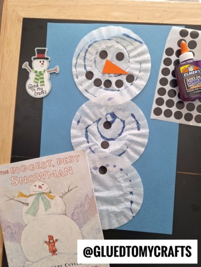 A snowman craft made with three coffee filters on blue paper, decorated with dot stickers and a paper carrot nose. Beside it are a snowman sticker, glue, dot stickers, and the book The Biggest, Best Snowman.