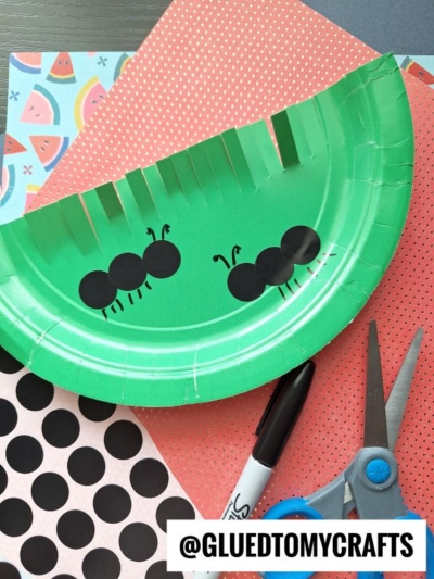 A green paper plate decorated as a watermelon slice with black dot stickers forming two ants, drawn legs and antennae, scissors, a marker, and patterned paper in the background.