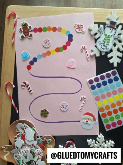 A pink paper with a curvy path traced in purple, decorated with colorful dot stickers, coding label crafts, and Christmas-themed stickers like Santa, candy canes, and snowflakes, surrounded by craft supplies and candy canes on a table.