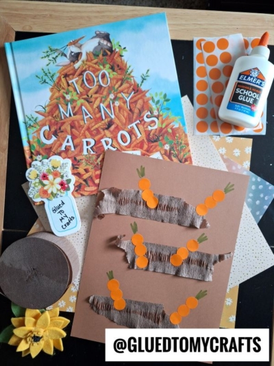 A children’s book titled “Too Many Carrots,” Elmer’s glue, patterned papers, a brown bunny craft with orange paper carrots, a felt flower, and a tag reading “Glued to My Crafts” displayed on a table.