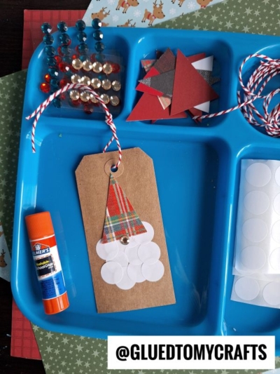 A blue tray holds red and white craft supplies, glue, gems, and a paper tag with a gnome made of plaid paper and circle stickers. Sticker sheets and a coding label crafts GLUEDTOMYCRAFTS label are visible.