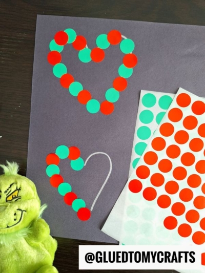 A piece of dark paper with two heart shapes outlined in white, each filled with red and green circle stickers—perfect for coding label crafts. Sticker sheets and a green plush toy are nearby. The text @GLUEDTOMYCRAFTS is at the bottom.