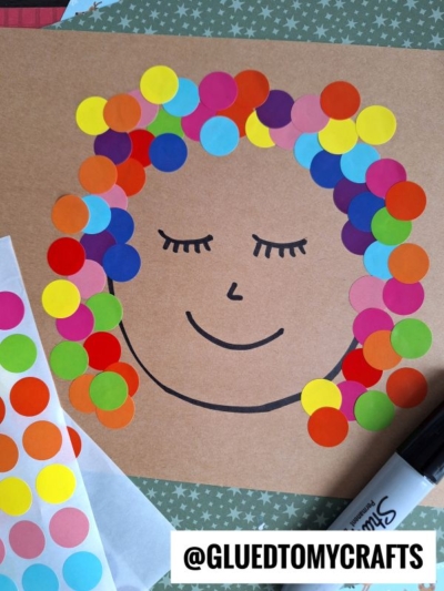 A drawing of a smiling face with closed eyes on brown paper, decorated with colorful round stickers as hair. Marker, sticker sheets, and a coding label reading “@GLUEDTOMYCRAFTS” capture the fun of creative crafts.