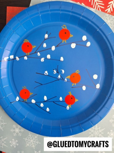 A blue paper plate decorated with red and orange paper circles as birds sits on painted brown branches with white snow dots. The birds have yellow beaks and legs, with a coding label crafts tag, @GLUEDTOMYCRAFTS, at the bottom.
