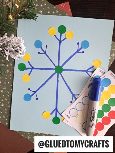 A blue paper features a snowflake drawn with a blue marker, decorated with colorful round stickers—a festive idea perfect for holiday label crafts. Nearby are a marker, sticker sheets, holiday paper, and a pine branch. @GLUEDTOMYCRAFTS is written at the bottom.