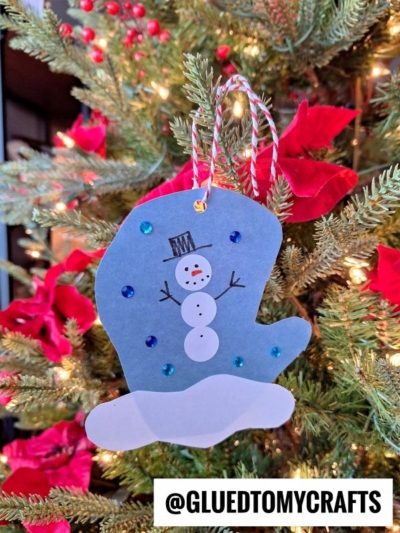 A blue mitten-shaped ornament decorated with a snowman, sequins, and white paper snow hangs on a Christmas tree with red poinsettias and lights—a charming addition to your coding label crafts. Text at the bottom reads @GLUEDTOMYCRAFTS.