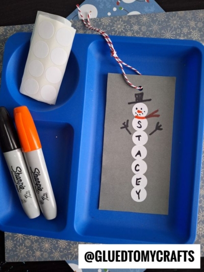 A blue tray holds black and orange Sharpie markers, a roll of round white stickers, and a coding label craft tag showing a snowman made from stickers spelling “STACEY.” The tag has a string and is labeled “@GLUEDTOMYCRAFTS.”.
