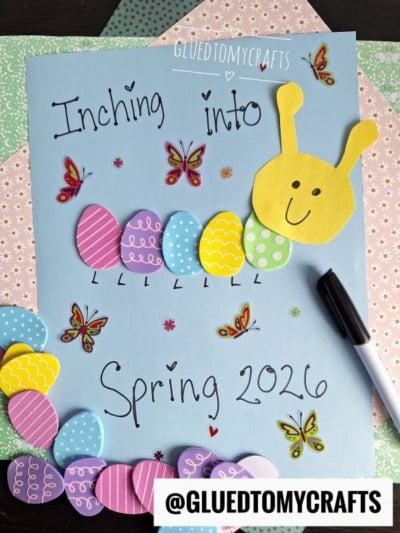 A spring craft featuring a blue paper with “Inching into Spring 2026” written on it, decorated with colorful paper eggs, butterflies, and a yellow paper caterpillar with a smiling face. A black marker lies beside the artwork.