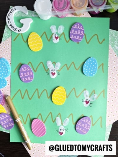 A green sheet of paper decorated with colorful paper Easter eggs, white bunny faces made from fingerprints, gold squiggly lines, and paint supplies nearby. @GLUEDTOMYCRAFTS is written at the bottom.