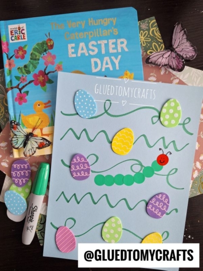 A childrens book titled The Very Hungry Caterpillars Easter Day is next to a blue craft sheet decorated with a caterpillar made of green circles, colorful paper Easter eggs, and wavy green lines. Two butterfly decorations, a Sharpie marker, and a @GLUEDTOMYCRAFTS label are also visible.