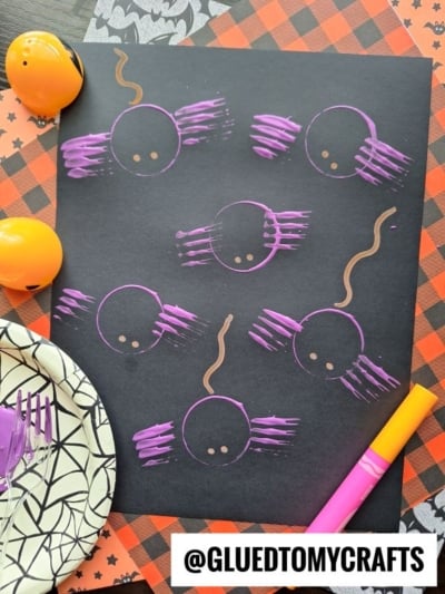 A black paper features six purple spider crafts made with fork prints and brown squiggly lines for webs, surrounded by Halloween-themed items like orange eggs, a spiderweb plate, purple fork, and orange marker.