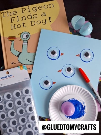 A childrens book, The Pigeon Finds a Hot Dog!, lies next to blue paper with googly eyes, a red marker, paint, and plastic eggs, set up for a craft project. @GLUEDTOMYCRAFTS is displayed in the corner.
