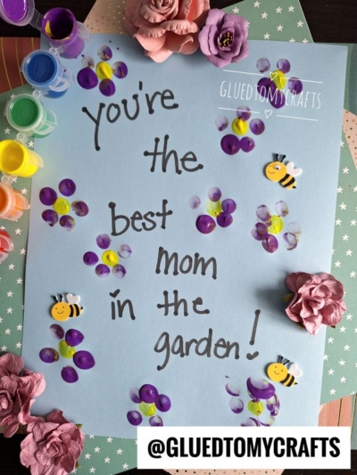 A blue paper reads Youre the best mom in the garden! surrounded by purple and yellow fingerprint flowers, bee stickers, paint pots, paper flowers, and has the handle @GLUETOMYCRAFTS at the bottom.