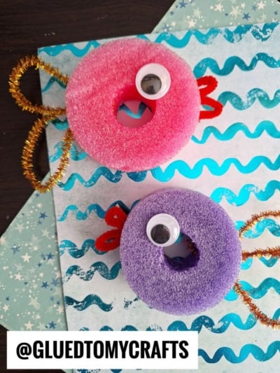 A pink and a purple foam ring each decorated with one googly eye, orange pipe cleaner antennae, and sitting on blue-patterned paper. Text at the bottom reads @GLUEDTOMYCRAFTS.