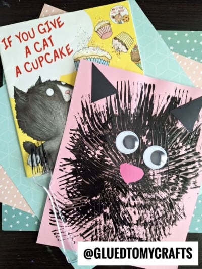 A children’s book titled If You Give a Cat a Cupcake and a pink fork painted cat craft with big googly eyes, black painted body, and pink paper ears and nose, displayed on a pastel patterned background. @GLUEDTOMYCRAFTS is written below.