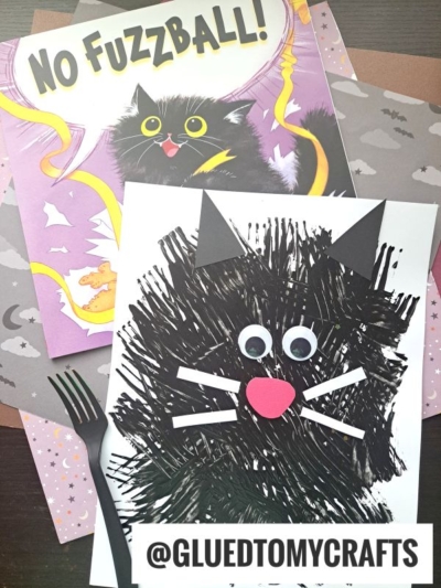 A children’s book titled “No Fuzzball!” with a black cat on the cover sits beside a fork painted cat craft, featuring googly eyes, white whiskers, and a pink nose, on top of colorful paper. @GLUEDTOMYCRAFTS is written at the bottom.