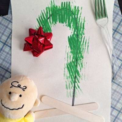 A simple Christmas tree made with green fork prints on paper, decorated with a red bow, craft sticks, and a plush Charlie Brown toy beside it. The handle @GLUEDTOMYCRAFTS is written at the bottom.