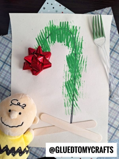A simple Christmas tree made with green fork prints on paper, decorated with a red bow, craft sticks, and a plush Charlie Brown toy beside it. The handle @GLUEDTOMYCRAFTS is written at the bottom.