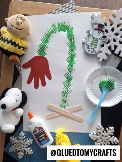 A holiday craft setup features a paper with a green painted Grinch arm, a red handprint for the Grinchs hand, craft sticks, Elmer’s glue, a plate with green paint, Snoopy and Charlie Brown plush toys, snowflake decorations, and a snowman ornament.