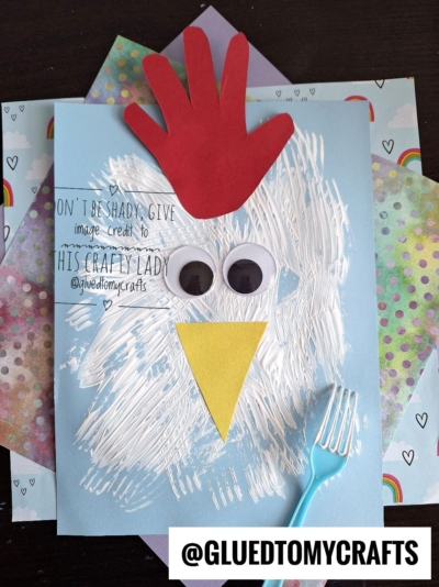 A childrens chicken craft made from a blue paper, white painted strokes, two large googly eyes, a yellow paper beak, a red handprint for a comb, and a blue plastic fork, displayed on rainbow-patterned sheets.