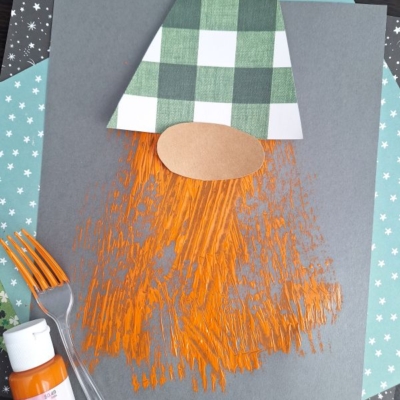 A gnome craft made from paper shapes with a green-checkered hat and orange painted beard, created using a plastic fork. Supplies include orange paint, a fork, and patterned paper. The handle @GLUEDTOMYCRAFTS is visible.