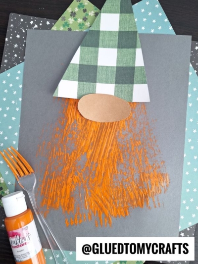 A gnome craft made from paper shapes with a green-checkered hat and orange painted beard, created using a plastic fork. Supplies include orange paint, a fork, and patterned paper. The handle @GLUEDTOMYCRAFTS is visible.