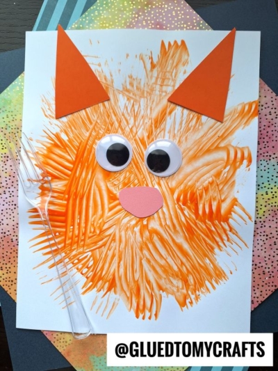 A cat craft made from orange paint streaks, large googly eyes, orange paper ears, and a pink paper nose on white paper, with a plastic fork and a colorful background. Text reads @GLUEDTOMYCRAFTS.