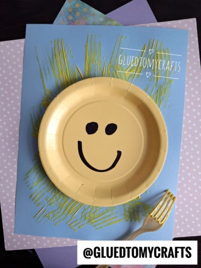 A yellow paper plate with a black smiley face is placed on blue paper decorated with yellow paint strokes, resembling a sun. The setup is on top of layered pastel paper sheets with patterns. A gold fork is beside the plate.