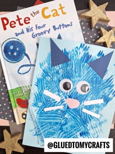A childrens book Pete the Cat and His Four Groovy Buttons lies next to a blue cat craft made with painted strokes, googly eyes, paper ears, whiskers, and a pink nose. Gold stars and a @GLUEDTOMYCRAFTS label are nearby.