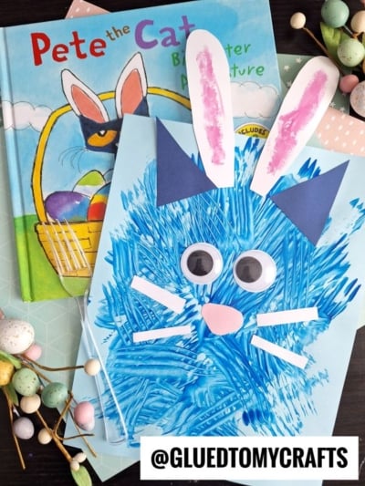 A blue textured craft bunny with paper ears, googly eyes, and whiskers is placed on children's books, including Pete the Cat. Beside the artwork are decorative pastel eggs, white berries, and a colorful fork painted cat.