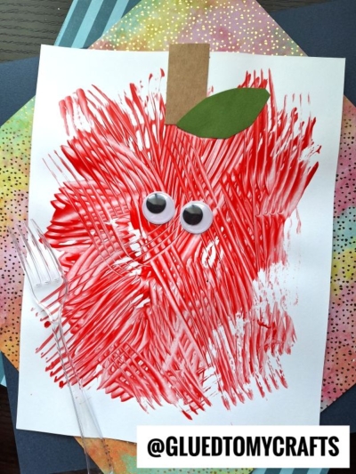 A child’s craft of a red apple made with fork paint streaks, featuring two large googly eyes and a green paper leaf on white paper—a fun idea for FALL CRAFTS FOR KIDS. The art is taped to colored paper, labeled @GLUEDTOMYCRAFTS at the bottom.