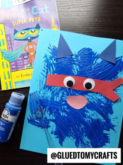 A children’s craft of Super Pete the Cat with blue painted fur, paper ears, a red mask, googly eyes, and a nose on blue paper. A Pete the Cat: Super Pete book, blue paint, and a plastic fork are nearby.