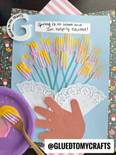 A spring-themed craft shows a bouquet of painted tulips made with a brown paper hand, doily, and colorful fork-stamped petals on blue paper. A sign reads, Spring is in bloom and Im tulip-ly excited!.