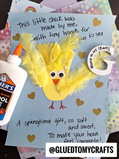 A handmade card features a yellow chick made of feathers and paper with googly eyes on blue paper, surrounded by gold hearts. Theres a glue bottle, craft supplies, and the Instagram handle @GLUEDTOMYCRAFTS.