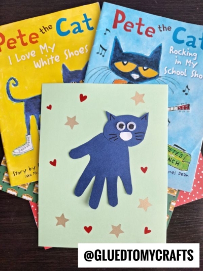 A handmade card with a blue cat made from a cutout handprint is placed on top of two Pete the Cat books. The card is decorated with star stickers and the Instagram handle @GLUEDTOMYCRAFTS is at the bottom.