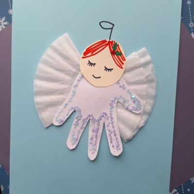 A child’s handprint cutout decorated as an angel, with a paper face, cupcake liner wings, red hair, and glitter, is glued to blue paper. The Instagram handle @GLUEDTOMYCRAFTS is at the bottom right.