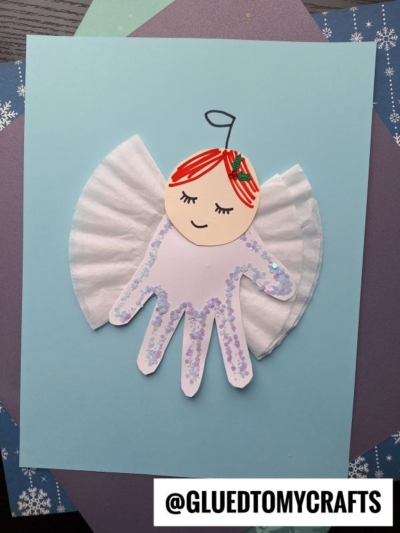A child’s handprint cutout decorated as an angel, with a paper face, cupcake liner wings, red hair, and glitter, is glued to blue paper. The Instagram handle @GLUEDTOMYCRAFTS is at the bottom right.