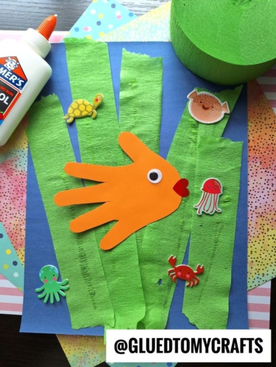 A kids’ underwater craft with green crepe paper seaweed, foam sea creature stickers, and an orange paper fish made from a handprint, all on a blue background. A bottle of glue and green crepe paper are nearby.