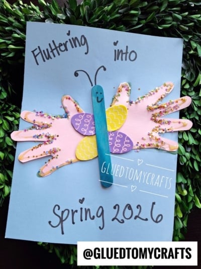 A handmade butterfly craft made from pink paper handprints, a blue popsicle stick, and colorful glitter, on a blue sheet with the words Fluttering into Spring 2026 and @GLUEDTOMYCRAFTS.