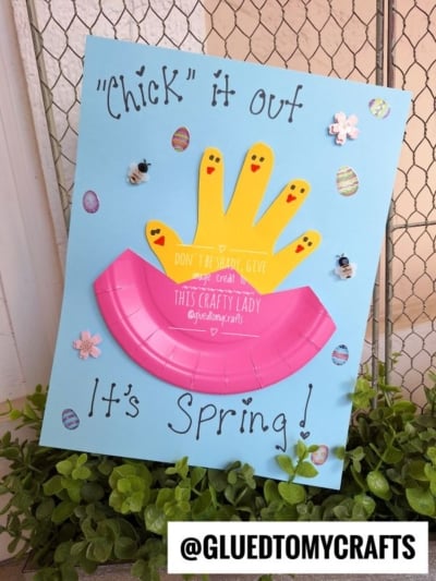 A spring craft on blue paper features three yellow handprint chicks, a pink paper plate, small bees, and decorated eggs. The text says Chick it out and Its Spring! with the handle @GLUEDTOMYCRAFTS at the bottom.