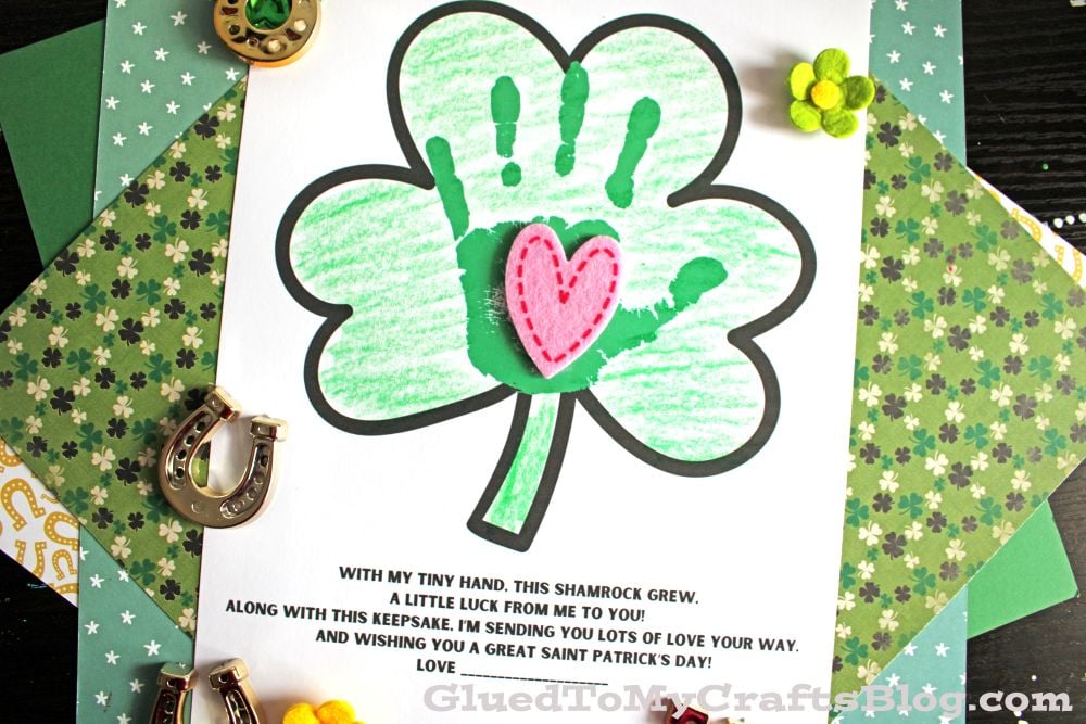 A St. Patrick’s Day craft features a shamrock colored green with a child’s handprint forming one leaf and a pink felt heart in the center. Surrounded by decorative horseshoes, flowers, and patterned paper, it pairs perfectly with St. Patrick's Day Printables.