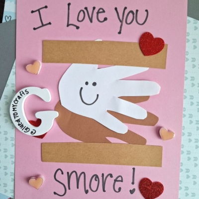 A pink handmade card features layered cutouts of handprints forming a s’more with a smiling face, surrounded by heart decorations. Text reads “I Love You S’more!” with the handle @GLUETOMYCRAFTS at the bottom.