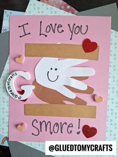 A pink handmade card features layered cutouts of handprints forming a s’more with a smiling face, surrounded by heart decorations. Text reads “I Love You S’more!” with the handle @GLUETOMYCRAFTS at the bottom.