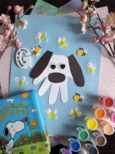 A children’s spring craft project features a dog made from a white hand cutout with black ears and googly eyes on blue paper, surrounded by bee stickers, paint, colored paper, blossoms, and a Snoopy book.