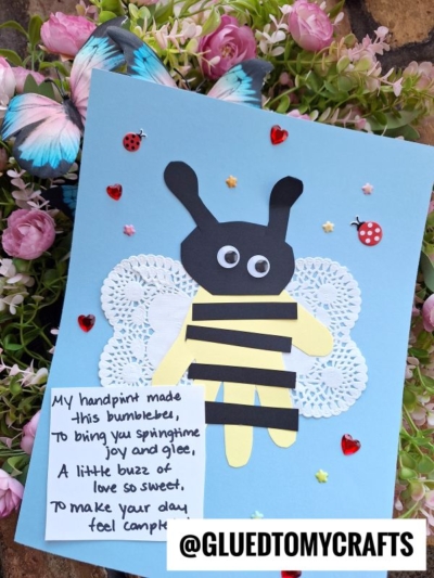 A handmade bumblebee craft on blue paper features a yellow handprint bee with black stripes and googly eyes, white doily wings, red heart and ladybug stickers, and a handwritten note. Flowers and butterflies surround it.