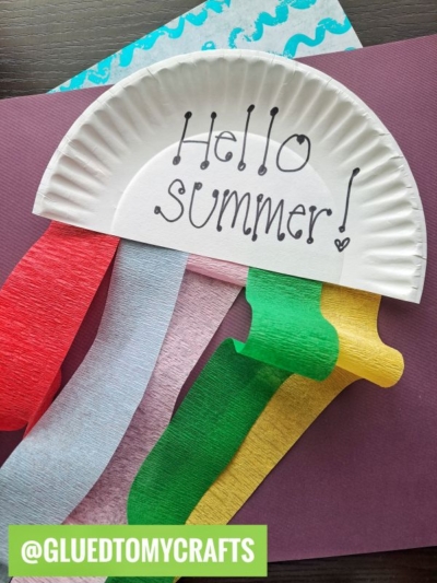 A paper plate cut in half with Hello Summer! written on it, decorated with wavy strips of red, white, green, and yellow paper, resembling a jellyfish, on colored paper backgrounds. Text: @GLUEDTOMYCRAFTS.